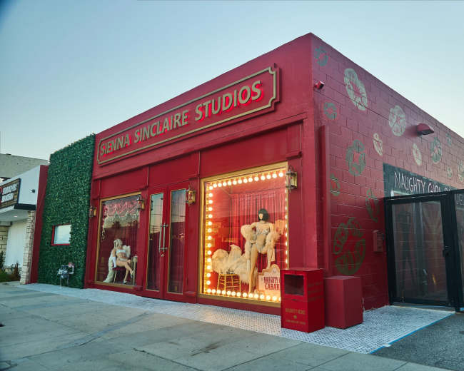 The image shows a red building with "Sienna Sinclaire Studios" written in bold letters, featuring large windows displaying mannequins and a gated entrance.