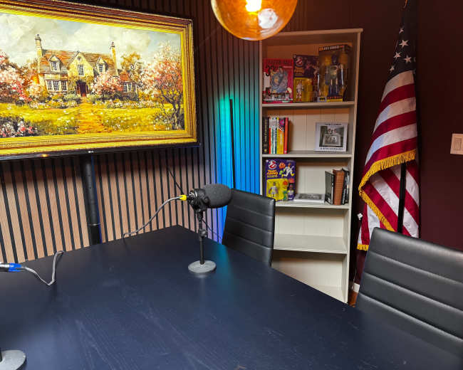 A dark table with two empty black chairs is set up in a room featuring a colorful painting, an American flag, and shelves filled with books and action figures.