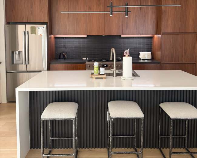 A modern kitchen features a central island with three stools, sleek cabinetry, and stainless steel appliances.