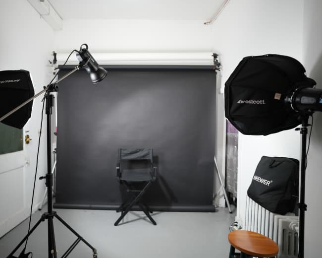 The image shows a photography studio setup with two softbox lights, a black backdrop, and a director's chair positioned in front of it.