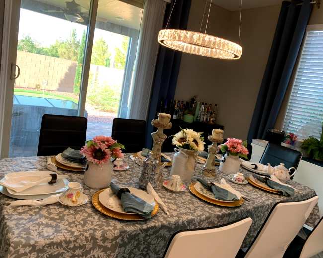 A dining table is set with decorative tableware, flowers in vases, and a chandelier overhead, all in a well-lit room with a large window overlooking a garden.