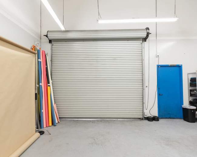 The image shows a storage area with a large rolled-up metal door, a blue door on the side, and several colorful backdrop sheets leaning against the wall.