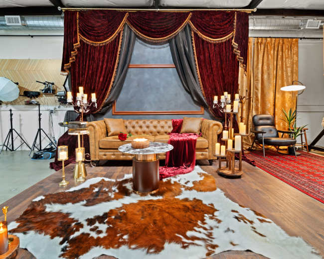 The scene features a decorated living space with a velvet sofa, a large cowhide rug, soft lighting from candles, and ornate curtains in the background.