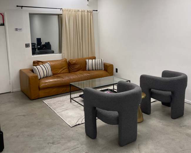 A simple room with a brown leather sofa, two gray armchairs, a glass coffee table, and a curtain covering a window.