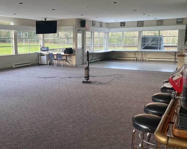 The image shows an empty room with large windows, a flat-screen TV mounted on the wall, and a vacuum cleaner on the floor, set up for a gathering or event.