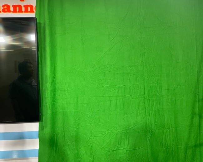 A green backdrop, commonly used for chroma keying, alongside a television screen and a person reflected in the glass.