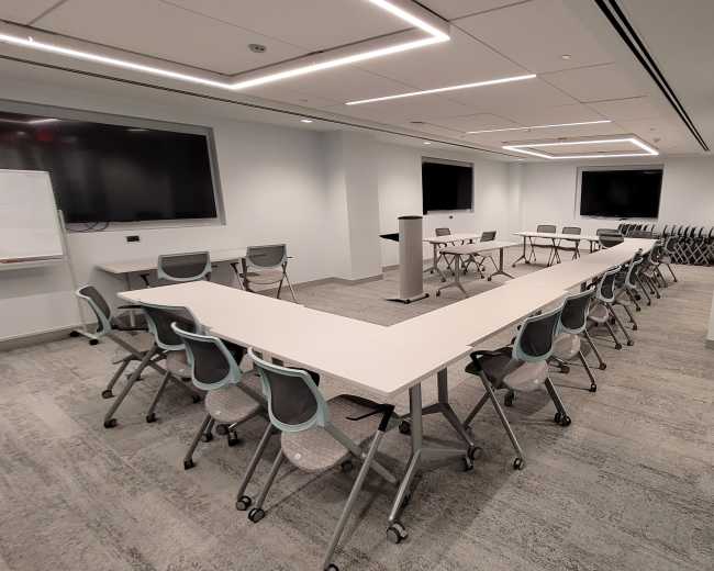 The image shows a modern conference room set up with a long table arranged in a U-shape, surrounded by chairs, with large screens mounted on the walls.