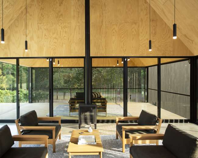 A modern living space features wooden walls, large glass windows, and minimalist furniture arranged around a central fireplace.