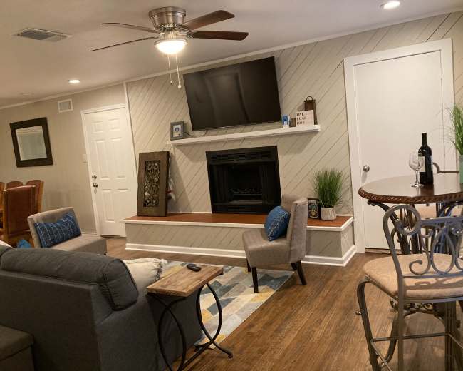 The image depicts a living room with a gray couch, two chairs, a coffee table, a fireplace with a television mounted above it, and a dining area in the background.