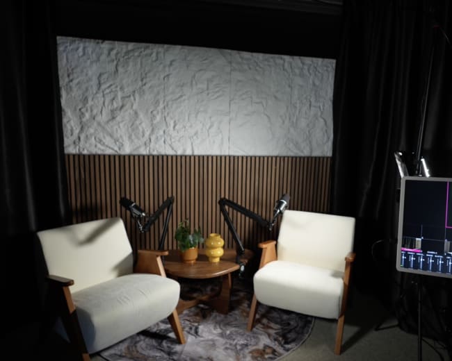 The image shows a well-lit indoor setting with two white chairs positioned across from a small round table, surrounded by acoustic treatment and a blank screen in the background.