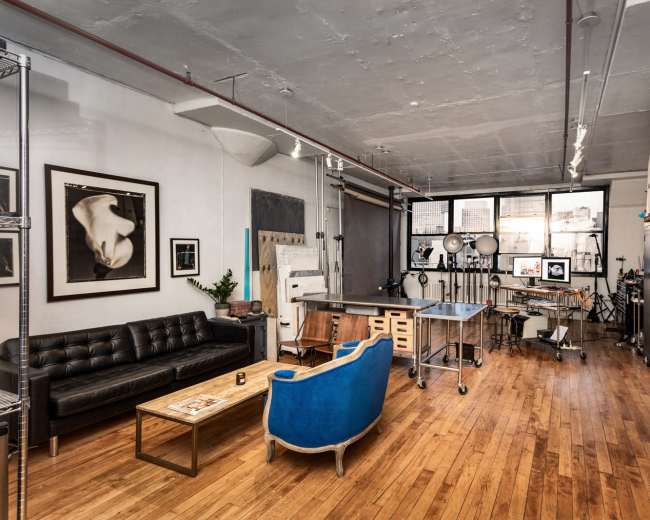 The image depicts a spacious studio with wooden floors, featuring a black leather sofa, a blue upholstered chair, and various art equipment along the walls.