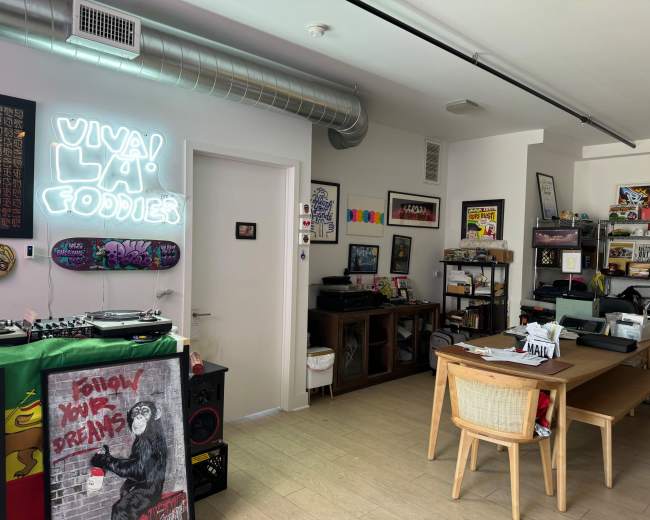 The image shows a room featuring a dining table, shelves filled with various items, a record player, and neon signs on the walls.