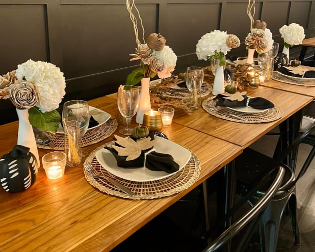 A long wooden dining table is elegantly set with plates, glasses, and decorative centerpieces featuring flowers and candles along a dark-paneled wall.