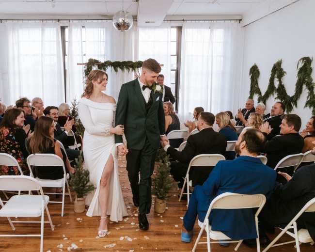 A bride and groom walk hand in hand down the aisle, surrounded by seated guests in a decorated venue.
