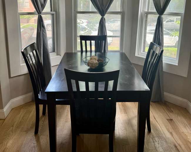 A dark wooden table is set in a well-lit corner with four chairs and large windows dressed with gray curtains.