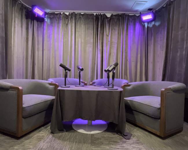 The image shows a small, cozy recording space with two armchairs, a table between them, and three microphones arranged on the table under purple lighting.