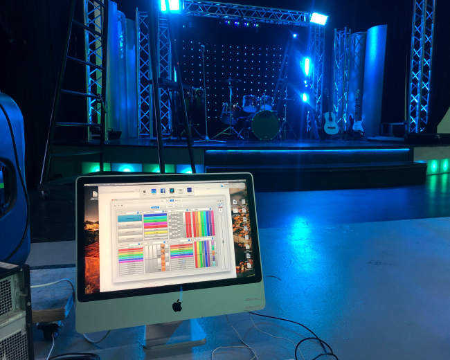 A computer displays software with a colorful interface in the foreground, while a stage set for a performance, featuring instruments and lighting, is visible in the background.