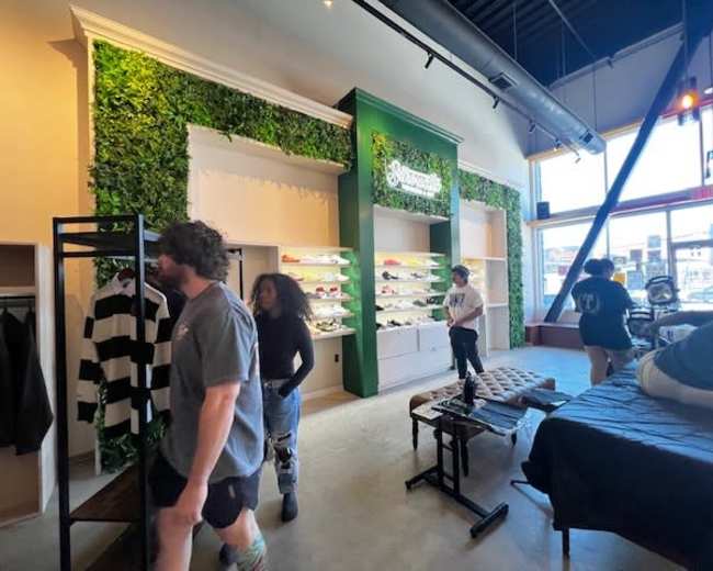 The image shows a clothing store interior with green foliage decor on the walls, clothing racks, and customers browsing the merchandise.