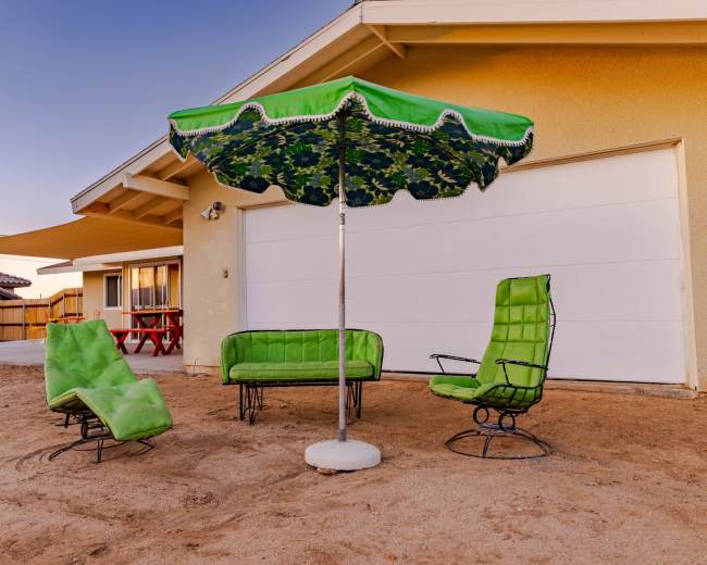 A set of green outdoor furniture, including two chairs, a sofa, and a large umbrella, is arranged on a sandy surface next to a garage.