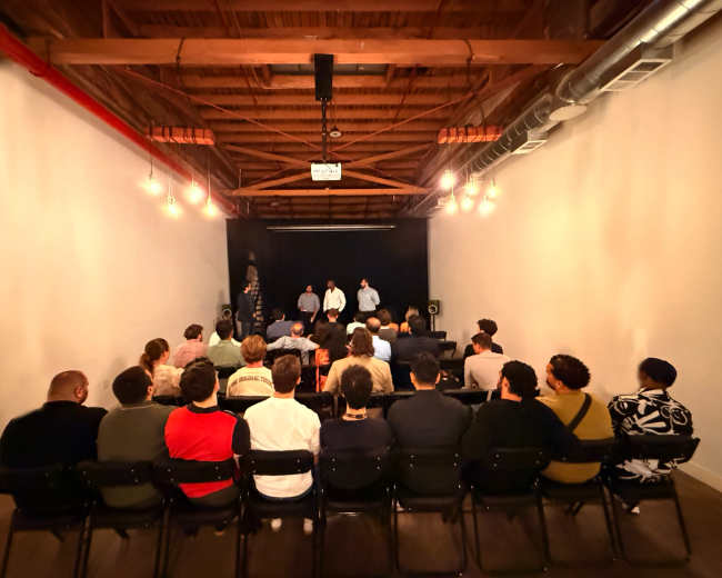 A group of people sits in chairs facing a small stage where two individuals are speaking, with warm lighting illuminating the scene.