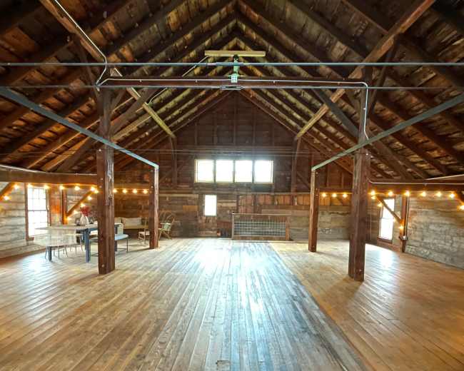 The image shows a large, rustic interior of an attic space with wooden beams, a high ceiling, and wooden flooring, illuminated by string lights.