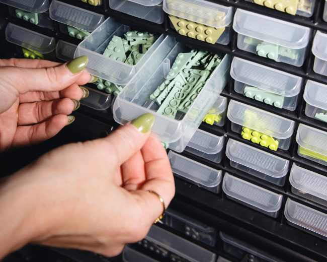 A person is opening two small plastic drawers filled with various green and yellow LEGO pieces.
