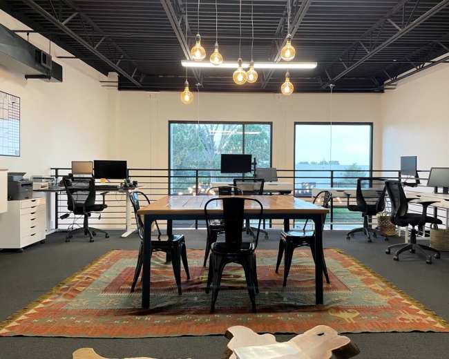 A modern office space features a large wooden table surrounded by chairs, with multiple computer workstations and large windows overlooking greenery.
