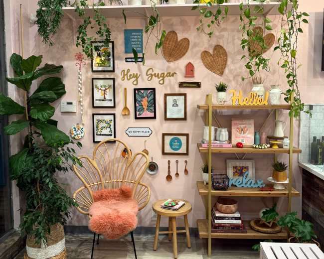 The image shows a cozy room corner featuring a decorative shelf filled with plants, framed art, and a wooden display unit, accompanied by a rattan chair and a small stool.