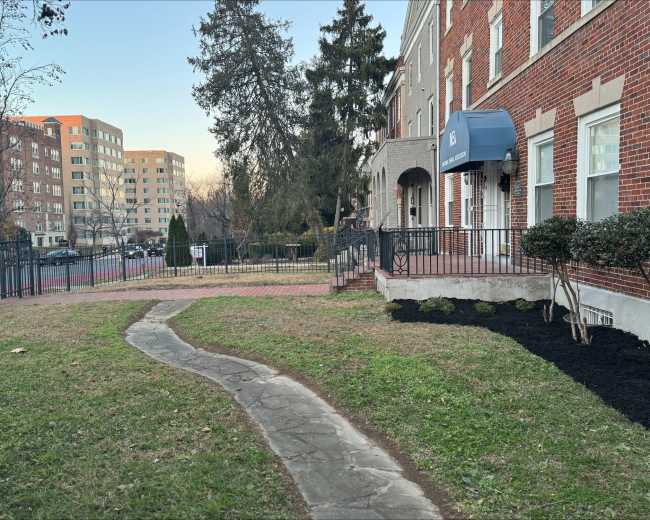 A winding stone pathway leads from a brick building with an awning to a grassy area, with trees and other buildings visible in the background.