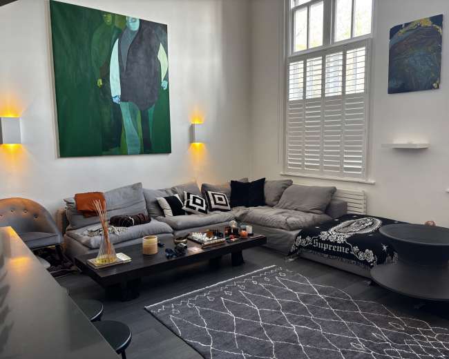 The image shows a modern living room with a gray sectional couch, a dark coffee table, and various decorative items, including artwork on the walls and a large area rug.