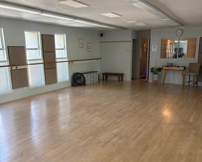 The image shows a spacious, empty room with wooden flooring, large windows, and a small kitchenette area at one end.