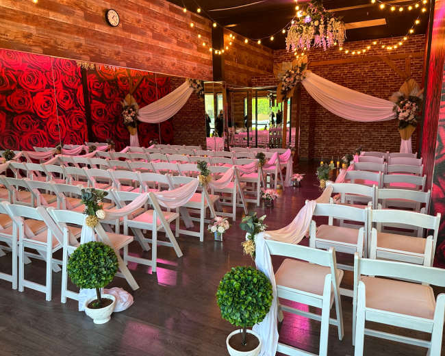 A decorated event space features rows of white chairs with floral accents, arranged facing a small altar, surrounded by rose-themed decor and soft lighting.