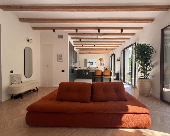 The image shows a modern living space with a large orange sofa in the center, wooden beams on the ceiling, and an open kitchen area in the background.