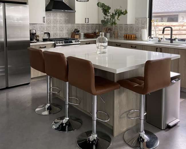 A modern kitchen features a large island with four brown bar stools, stainless steel appliances, and decorative pendant lighting above.