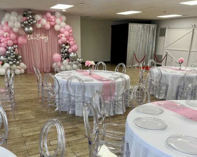 The image shows a decorated event space with round tables set with white tablecloths and transparent chairs, featuring a balloon backdrop and a pink curtain.