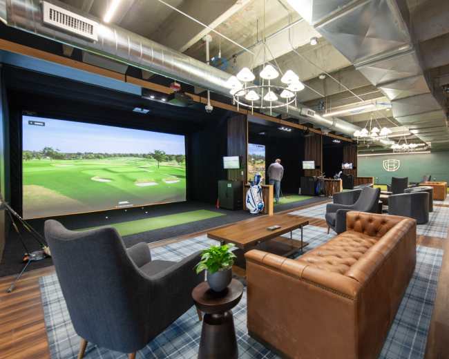 The image shows an indoor golfing lounge with a simulated golf screen, seating areas, and various golfing equipment.