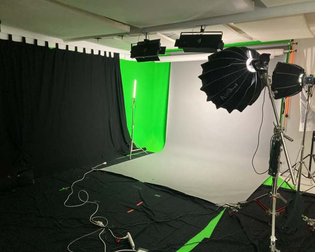 The image shows a photography studio setup with a green screen, a gray backdrop, and various lighting equipment arranged on the floor.