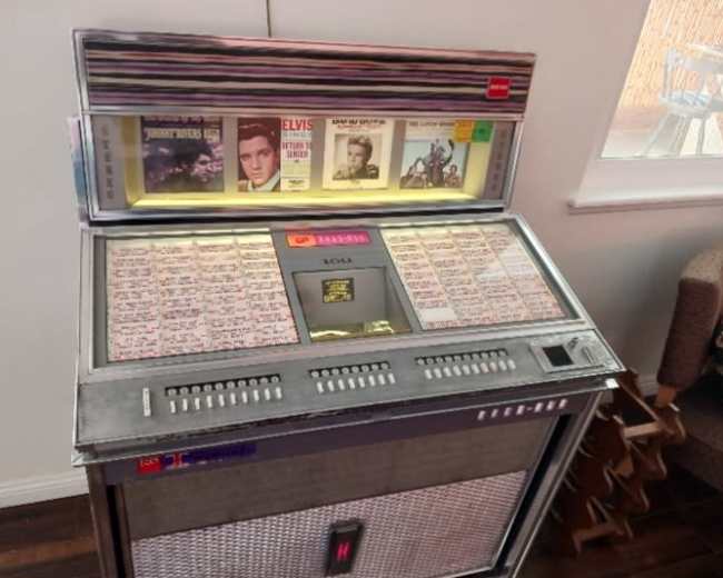 The image shows a vintage jukebox with vinyl records and a bright display, set against a light wall in an indoor space.