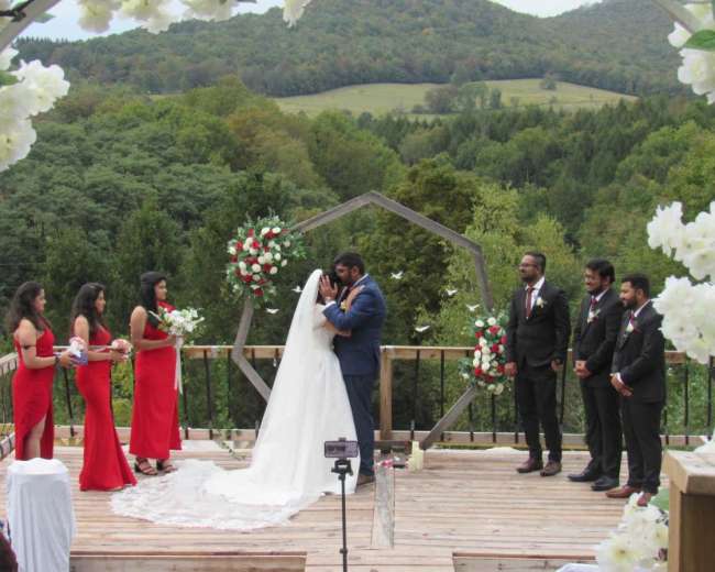 A bride and groom kiss under an arch adorned with flowers on a wooden deck surrounded by a lush green landscape.