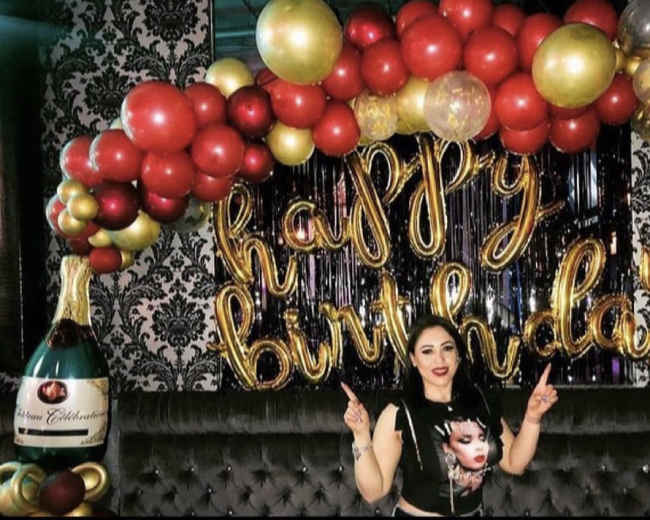 A woman poses in front of a decorated backdrop with balloons and a sign that reads "happy birthday."