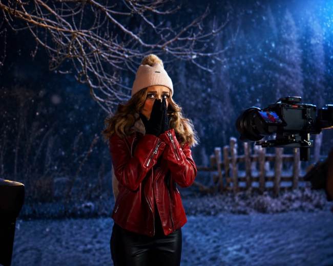 A woman wearing a red jacket and a white beanie stands outdoors in the snow, covering her face with her hands while a camera is directed at her.