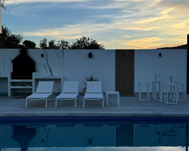 A swimming pool reflects the evening sky, surrounded by white lounge chairs and an outdoor dining area.