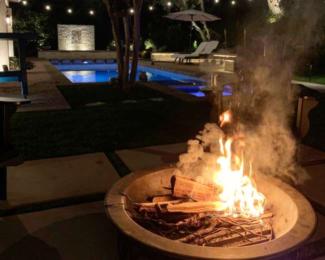 A backyard scene at night features a fire pit with flames, surrounded by stone pathways, a swimming pool, and softly lit trees.