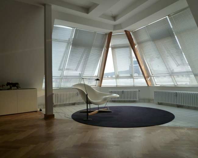 A spacious modern room with large windows, wooden structural elements, and a minimalist white chair on a circular dark rug.
