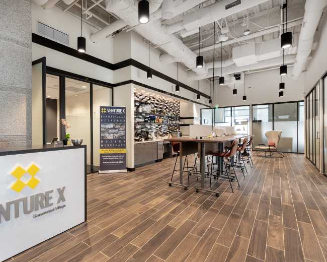 A modern coworking space featuring a reception area with a desk, a coffee station, high tables with stools, and glass-walled meeting rooms.