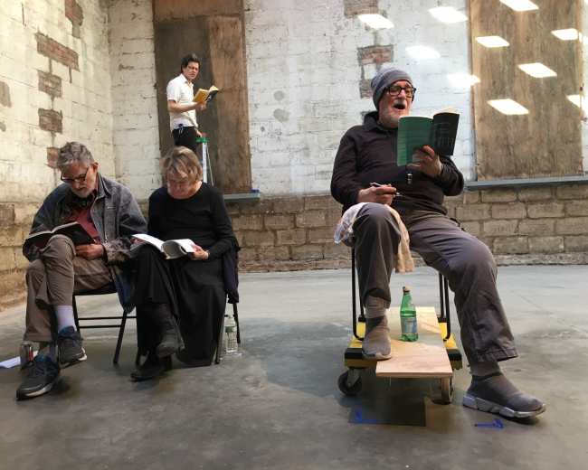 A group of four people is reading in an empty room with brick walls and scattered light coming through the upper windows.