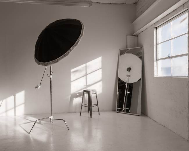 A photography studio features a large softbox, a mirrored wall, a stool, and sunlight casting shadows on the floor.