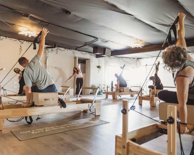 A group of people participates in a Pilates class using reformer machines in a well-lit studio.