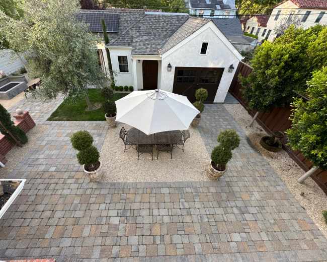 The image shows a well-designed patio area featuring a round table with chairs under a large umbrella, surrounded by decorative shrubs and a gravel pathway.