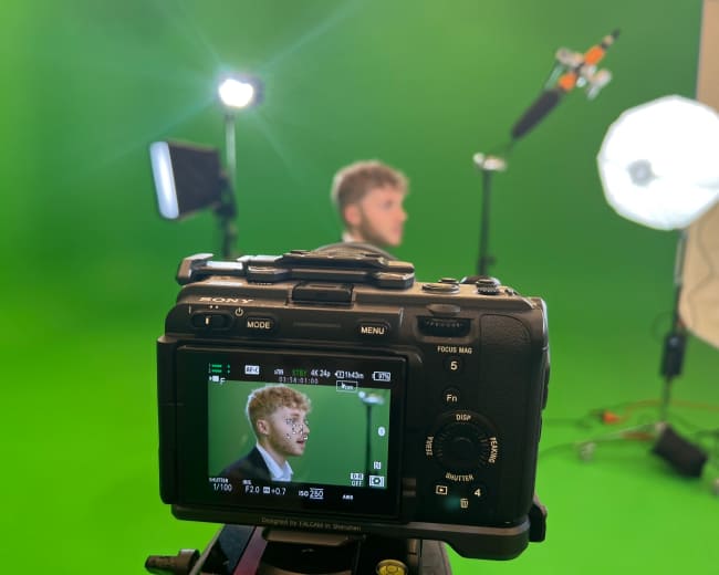 A camera is positioned on a tripod with its screen displaying a man in front of a green screen, while studio lights illuminate the scene.